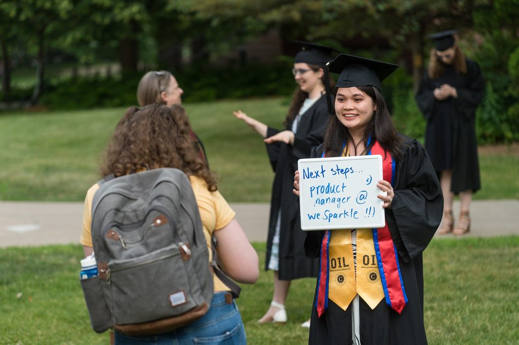 Class of 2023 Carleton student in graduation attire holding a sign saying their post-grad plans