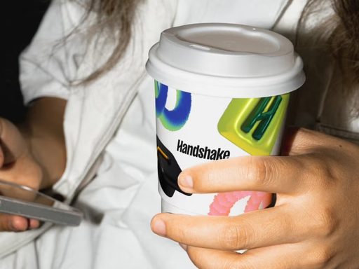 A young person holds a coffee cup with a logo for a company called handshake