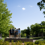 Five members of the Class of 2022, viewed from the back, stand on the Carleton College sign