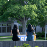 Two members of the Class of 2022, viewed from behind, hold up the 2022 banner while sitting on the Carleton sign