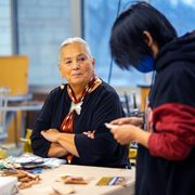 Dr. Denise Lajimodiere, 2022 Elder-in-Residence, sits at a table during her workshop on birch bark-biting. A student in the foreground is looking down at their piece of bark.