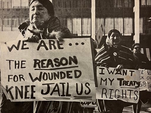 Black and white photograph of Native American elder women holding signs in protest on the anniversary of the 1973 occupation movement. Signs read: 