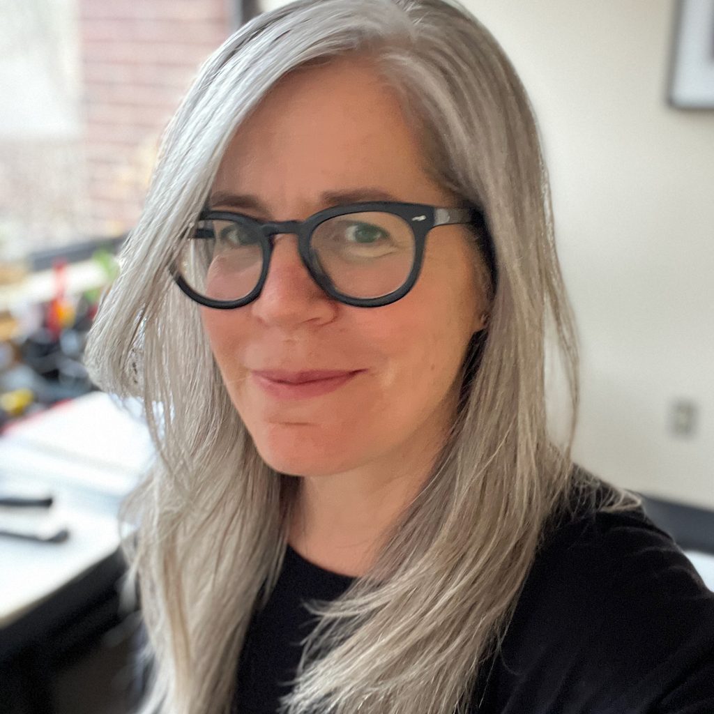 an image of a woman with grey hair and black glasses who is smiling at the camera