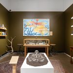 Inside a small room with olive green walls is a skeleton on a raised platform covered with dirt, an image of a bird and an octopus, a map on the back wall, a display case with artifacts, and many shelves covered with artifacts.