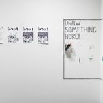 Four black-and-white comic book pages on a wall with a corner that says “Draw Something Here!” with markers and pens provided, upon which visitors have doodled.