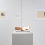 An open book on a pillar, with two black-and-white prints (pages from the book) mounted on the wall to either side.