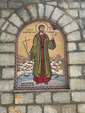 A mosaic depicting (Jesus?) in green and red robes, holding a cross, with a gold background and a port town (presumably Hydra) behind