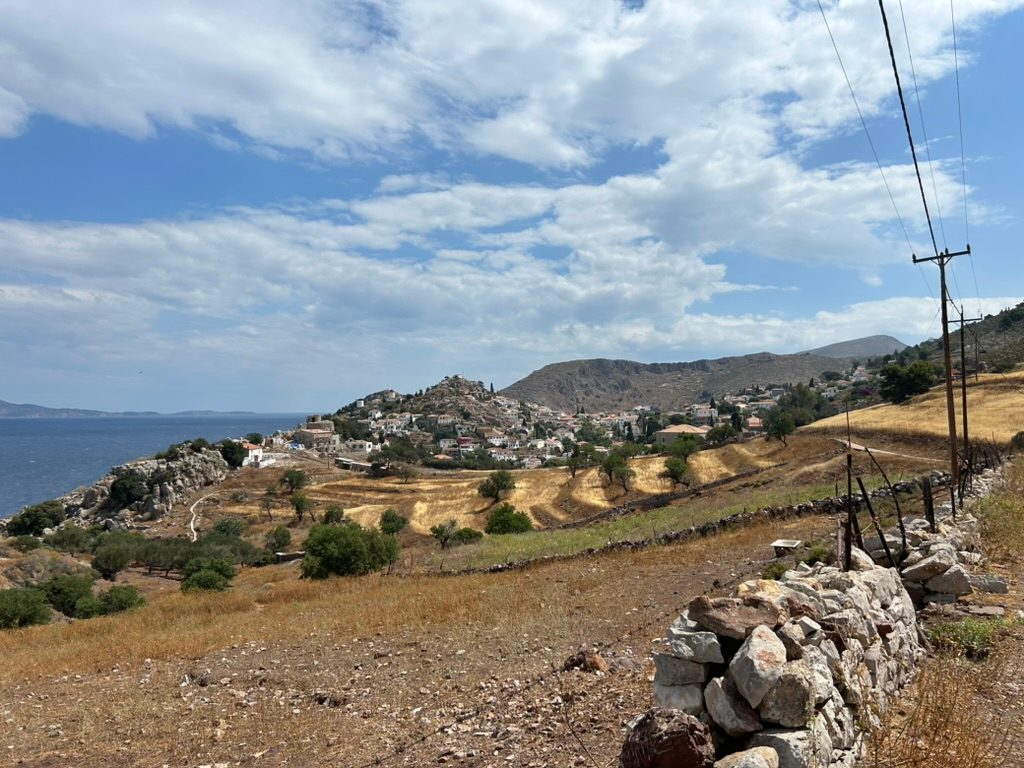 View of the town of Hydra over hills with fields and shrubs, from a path off the coastline. The town is visible beneath a blue sky with white clouds, and the photo is taken from behind a crude stone wall