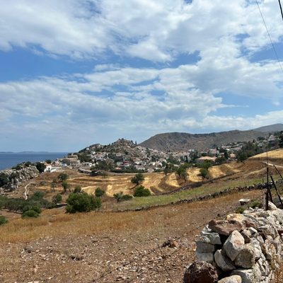 View of the town of Hydra over hills with fields and shrubs, from a path off the coastline. The town is visible beneath a blue sky with white clouds, and the photo is taken from behind a crude stone wall
