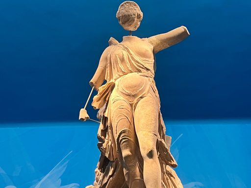 Statue of Nike in front of a blue museum background. Her arms, wings, and face are broken off.