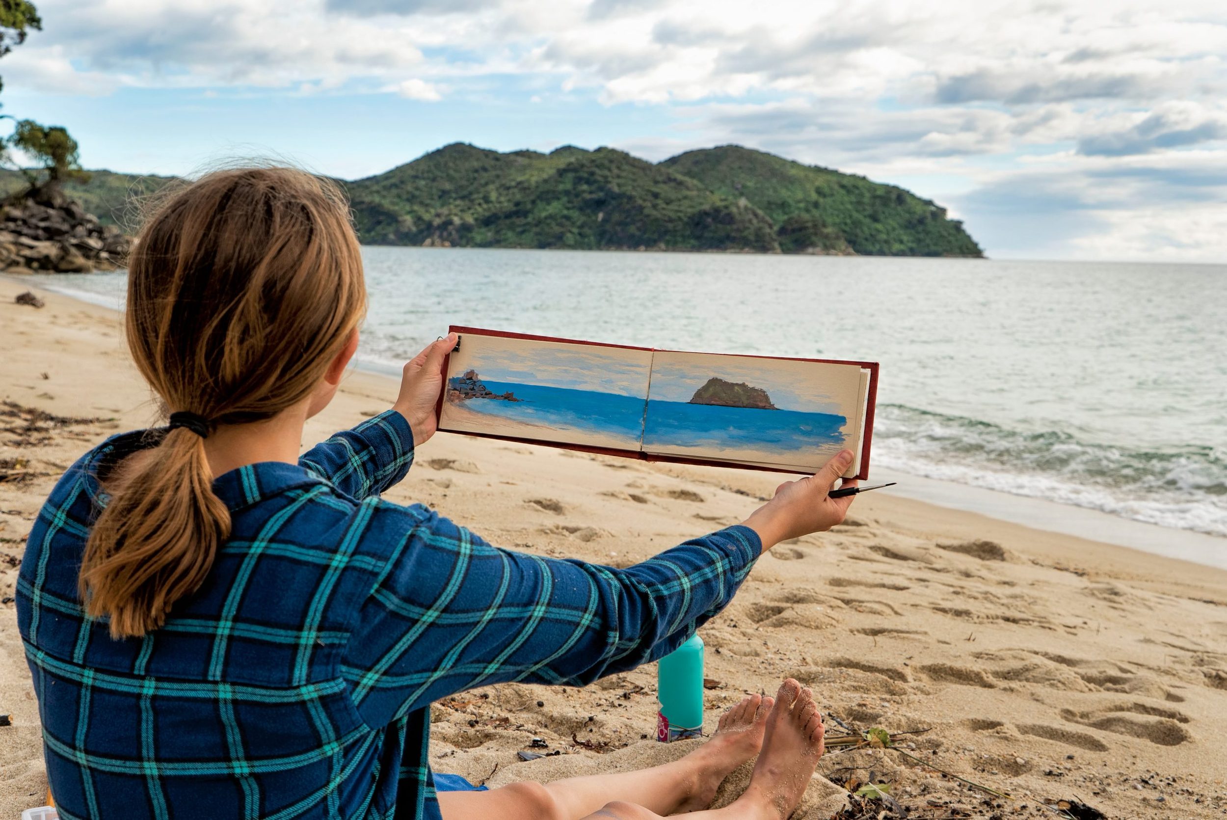 An artist holds their sketchbook up showing the landscape and the watercolor they created of it