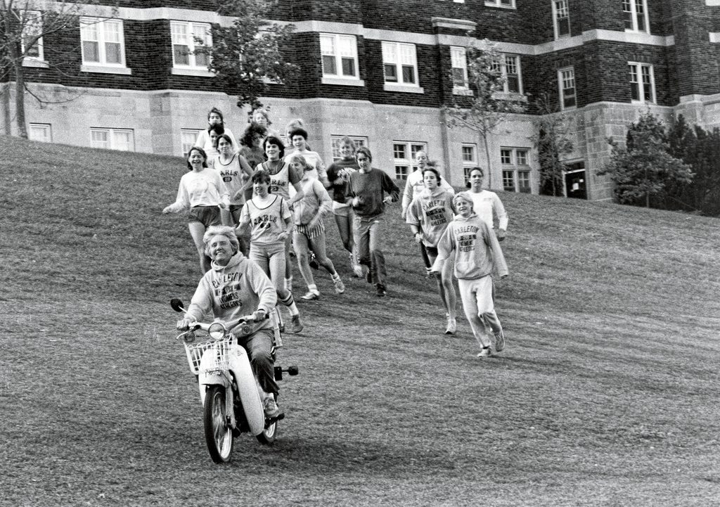 Ele Hansen rides down a hill on a scooter with a group of female students running behind her