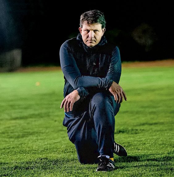 Coach Pete Sickle kneels on the grass of a rugby field