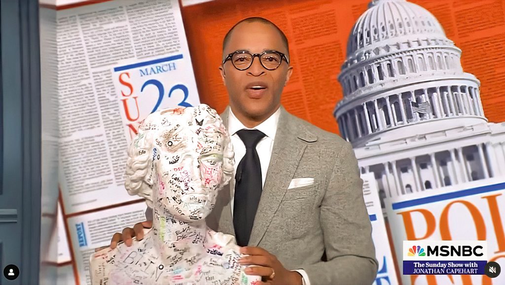 jonathan capehart poses at news desk with bust of schiller