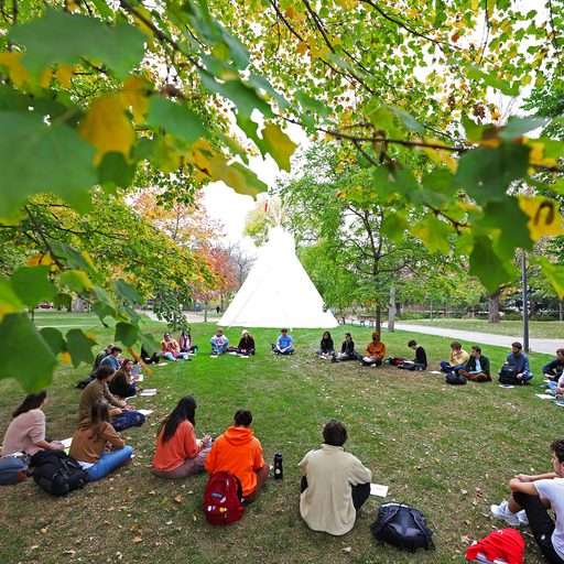 Students sit in a circle on campus with a Native American tipi in the background