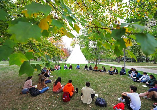 Students sit in a circle on campus with a Native American tipi in the background