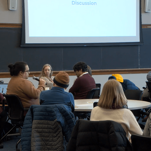 shows a group of people sitting around two round tables in a classroom, engaged in a discussion