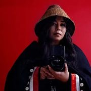 A portrait of Matika Wilbur. She is a Swinomish and Tulalip photographer wearing a traditional woven cedar hat and a dark blanket with a red and white stripe. She is holding a camera in front of her. The background is a solid red color.