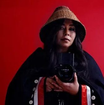 A portrait of Matika Wilbur. She is a Swinomish and Tulalip photographer wearing a traditional woven cedar hat and a dark blanket with a red and white stripe. She is holding a camera in front of her. The background is a solid red color.