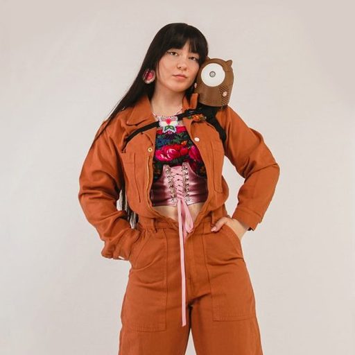 A full-body studio portrait of a young woman, Danielle Boyer, standing against a plain light background. She has long dark hair and is wearing a rust-orange jumpsuit over a pink lace-up corset and a brightly embroidered top. Her hands are in the jumpsuit pockets. An owl-shaped plush backpack is resting on her right shoulder. She is looking directly at the camera with a calm expression.