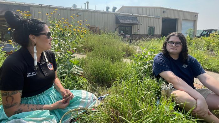 An Indigenous woman wearing a turquoise ribbon skirt and a black polo shirt labeled "Land and Environment" sits in a field of tall grass and yellow wildflowers. She is looking toward a person with long curly hair and glasses who is sitting cross-legged across from her. In the background, there is a large tan industrial-style building under a bright, clear sky.
