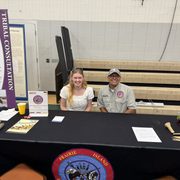 A young woman with long blonde hair and a man in a grey baseball cap sit behind a table at a Prairie Island Indian Community event. The table, draped in a black cloth featuring a large tribal logo, displays various archaeological artifacts—including stone tools and bones—alongside informational signs. To the left, a tall purple banner is titled 