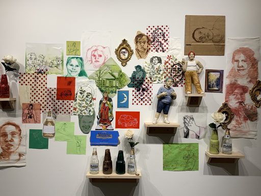 A wall collage of prints and drawings depicting different people in varying styles sit behind five shelves holding actual and sculpted beer bottles and ceramic sculptures of figures