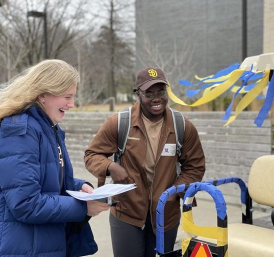 Two students laughing while standing next to a golf cart decorated with blue and yellow streamers.
