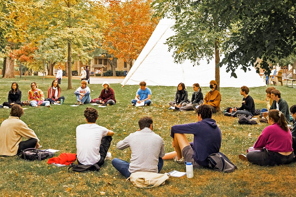 A class meets outside sitting in a circle, with a native American tipi in the background