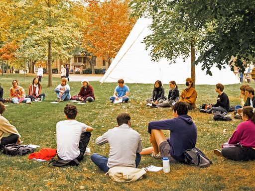 A class meets outside sitting in a circle, with a native American tipi in the background
