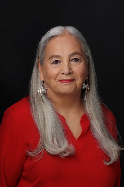 A studio portrait of Denise Lajimodiere, an Indigenous woman with long, straight silver-gray hair falling over her shoulders. She is wearing a red v-neck top, silver dragonfly drop earrings, and soft red lipstick, set against a dark background.
