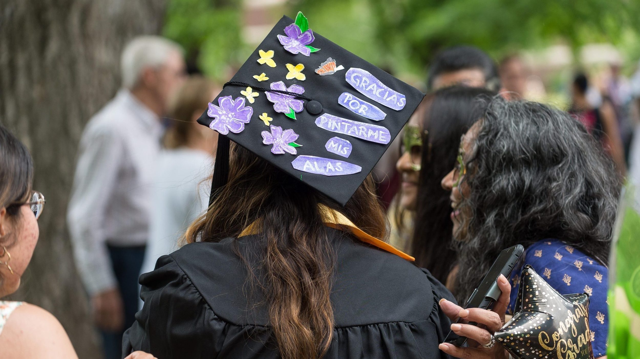 A student at commencement with her family. A message on her mortar board cap reads "gracias por pintarme mis alas"