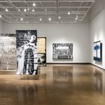 A brightly lit, white-walled gallery with concrete floors and high ceilings. Four very large-scale woven tapestries hang suspended from the lighting grid, two are mounted on the walls, and an artists’ book in a protective plexi-glass case appears on the right hand side of the image.