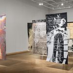 Five tapestries, approximately eight ft each tall hover between the floor and lighting grid, casting angular shadows across the museum floor. Staggered into three rows, design elements include typed and handwritten correspondence, a black and white image of two men under a tree, an inverted black and white image of a man wielding a gun.