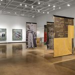 A rear view of five large woven tapestries, suspended from the lighting grid. On the wall in the far back hang two square-sized tapestries with green foliage on the perimeter and figures in the center.