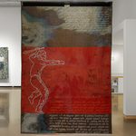 A rear view of a woven tapestry, approximately 8 ft tall. It is sectioned into three parts with the middle depicting a white outline of a figure weilding a baseball inside a blood red rectangular band of color. Above and below are areas of white text against a blended blue, white and brown background.