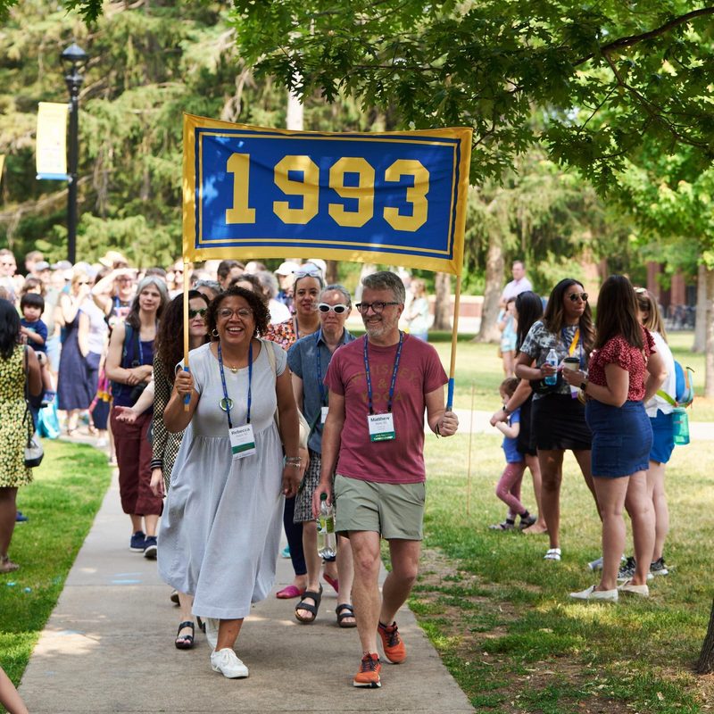 A man and a woman together hold a banner that says 1993 while walking down a sidewalk.