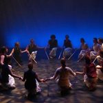 Dancers kneeling in a circle, each holding a stick
