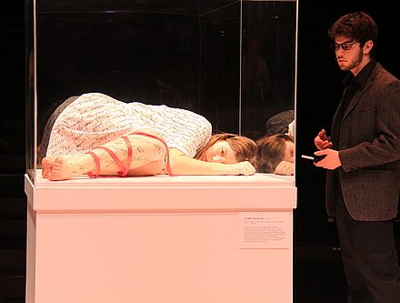 A woman laying in a museum display case, and a man looking at her
