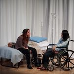 Woman sitting on a hospital bed, talking to a woman sitting in a wheelchair