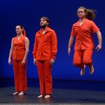 Dancers on stage wearing orange, one dancer jumping