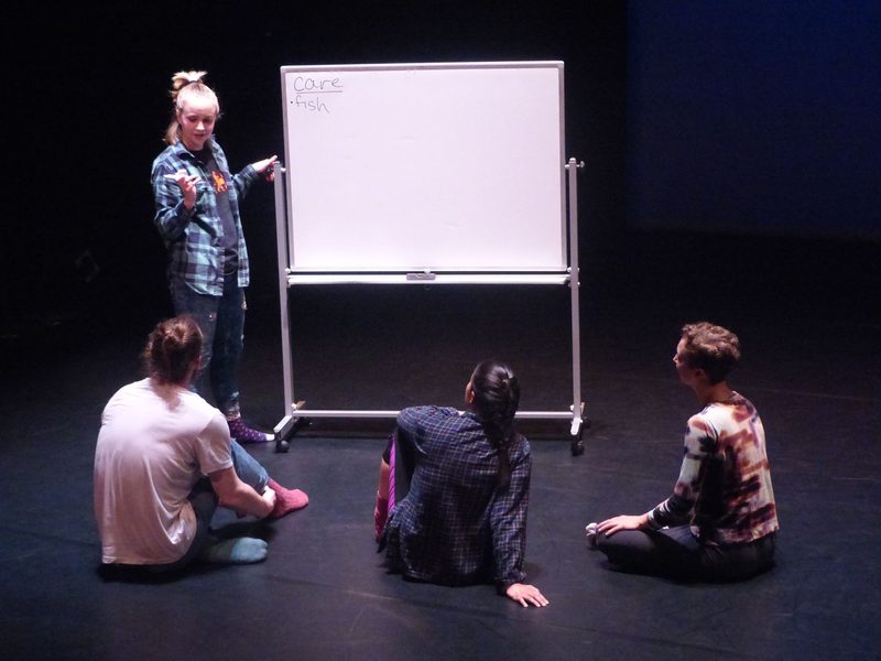 Dancer writing on a dry-erase board and three dancers seated in front of the board