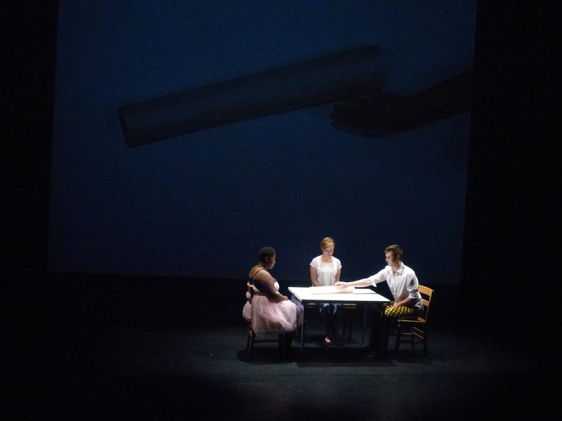 Dancers seated at a table, passing a roll of paper across the table and a projection of the paper