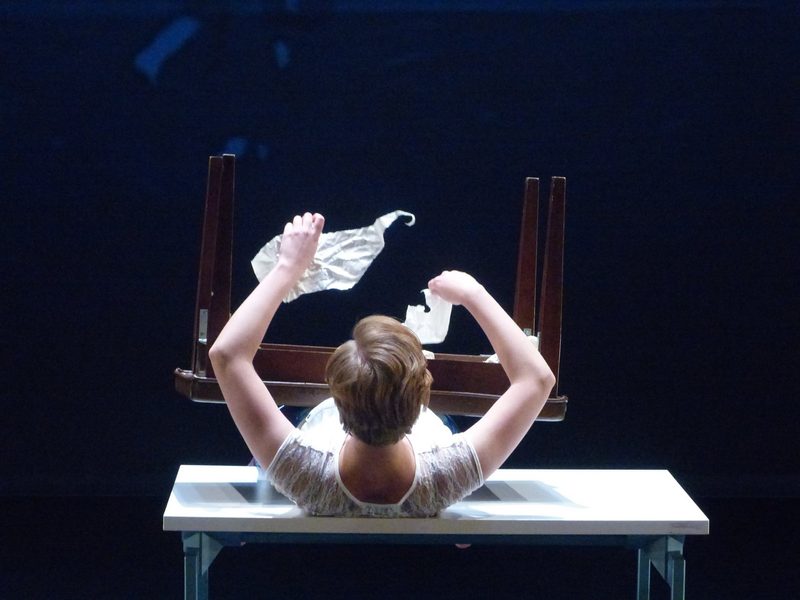 Dancer laying on a table, holding ripped paper, and a piano bench resting on top of her