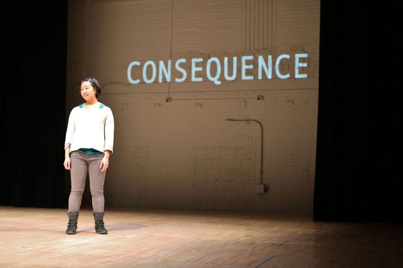 Girl standing on stage and the word "consequence" projected behind her