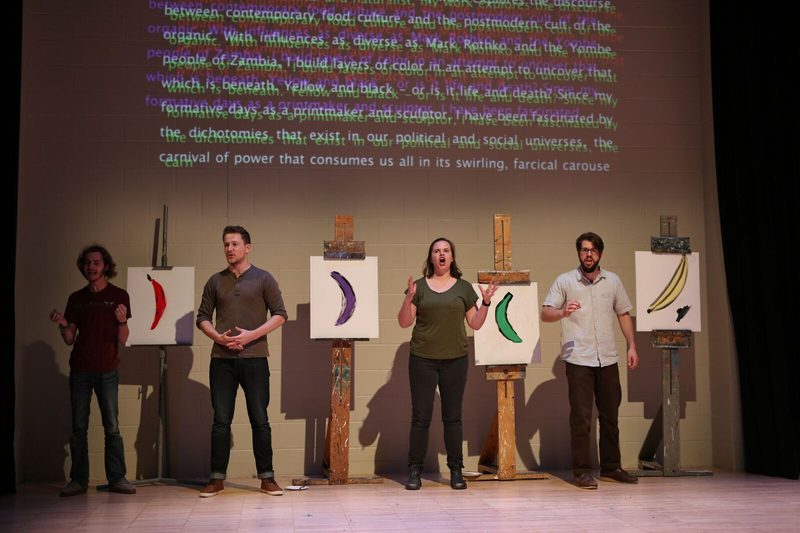 Actors standing next to paintings of colored bananas with words projected behind them