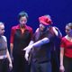 Four dancers standing next to a man with a red hat and his arm reached out.