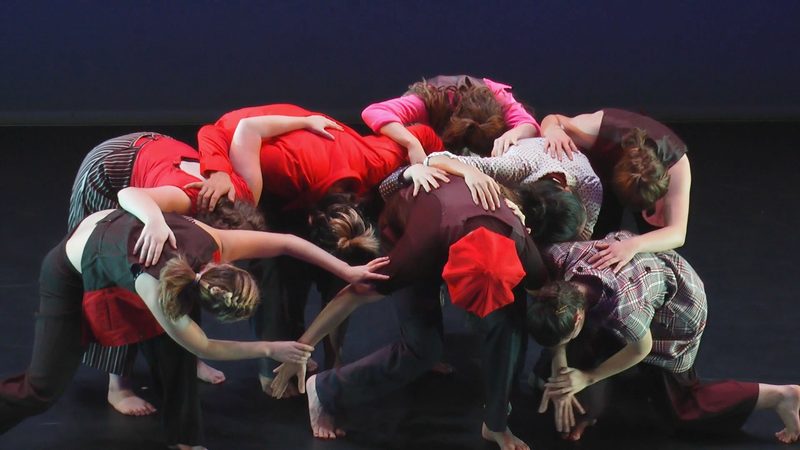 Dancers huddled together touching a dancer with a red hat in the middle.
