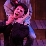 2 men fight onstage. One man holds the other in a choke-hold and he is gasping for breath.