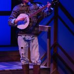 A man sings and plays banjo. He wears galoshes, ripped jeans, and a sweater.